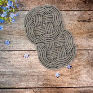 May include: Two gray woven round coasters with a knot design. The coasters are on a wooden surface.