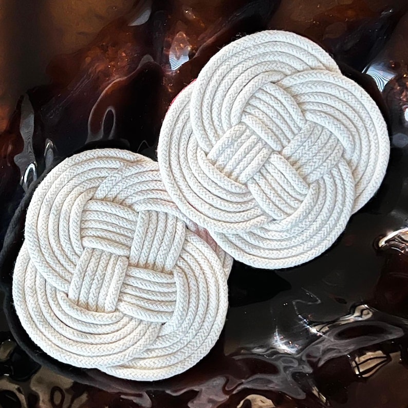 May include: Two white cotton rope coasters with a knotted design. The coasters are round and have a raised center.