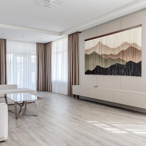 May include: A modern living room with a large wall hanging featuring abstract mountain shapes in shades of brown, green, and black. The wall hanging is made of yarn and is hung on a light brown wall. The room has white curtains and a light wood floor.
