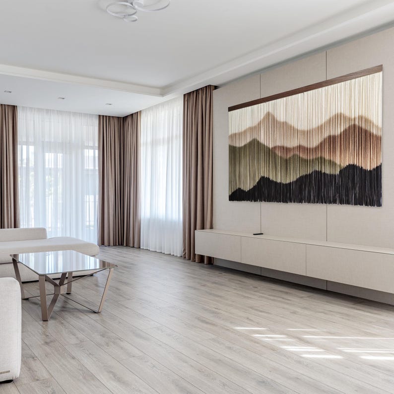 May include: A modern living room with a large wall hanging featuring abstract mountain shapes in shades of brown, green, and black. The wall hanging is made of yarn and is hung on a light brown wall. The room has white curtains and a light wood floor.