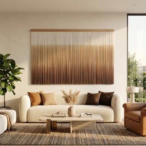 May include: A large rectangular wall hanging featuring vertical strands of fiber that transition in color from cream at the top to a deep brown at the bottom, suspended from a horizontal wooden rod mounted against a plain wall.