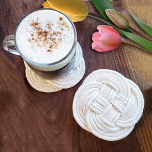 May include: Two white rope coasters with a knotted design. A glass of coffee with a layer of foam and cinnamon sprinkled on top sits on one of the coasters.