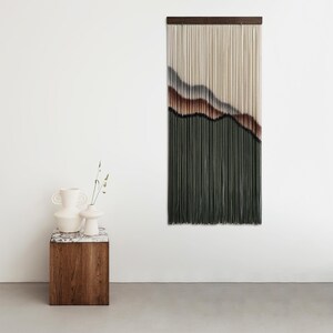 May include: A wall hanging made of yarn with a landscape design in shades of brown, grey, and green. The hanging is mounted on a wooden dowel rod. Two white ceramic vases are on a wooden table in front of the hanging.