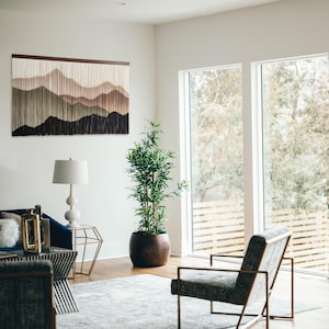 May include: A modern living room with a large window, a blue sofa, a side table, a lamp, a potted plant, and a chair. The wall features a macrame wall hanging with a mountain range design in shades of brown, green, and white.
