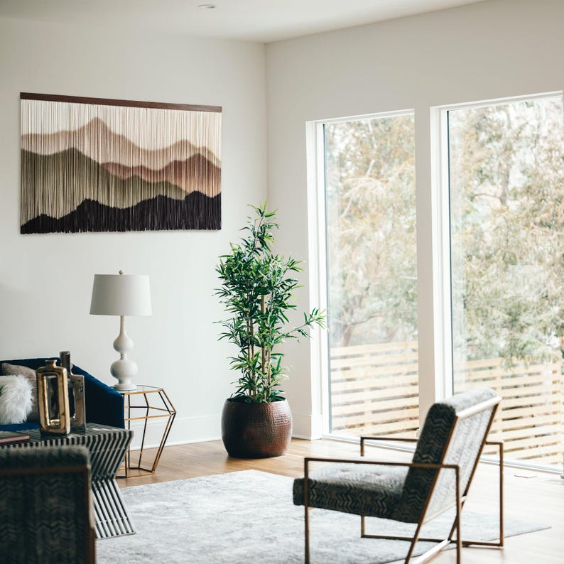 May include: A modern living room with a large window, a blue sofa, a side table, a lamp, a potted plant, and a chair. The wall features a macrame wall hanging with a mountain range design in shades of brown, green, and white.
