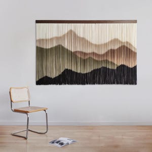 May include: A wall hanging featuring a woven tapestry with a landscape design. The tapestry is made of yarn in shades of brown, beige, green, and black. The tapestry is hung on a white wall with a wooden frame.