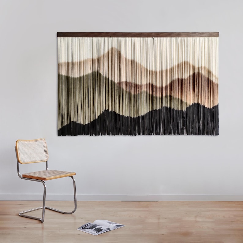 May include: A wall hanging featuring a woven tapestry with a landscape design. The tapestry is made of yarn in shades of brown, beige, green, and black. The tapestry is hung on a white wall with a wooden frame.