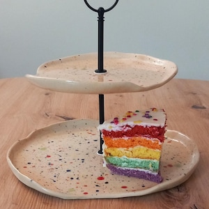 Könnte beinhalten: Ein zweistöckiger Servierständer mit schwarzem Griff und gesprenkelten beigefarbenen Tellern. Ein Stück Regenbogenkuchen mit weißem Zuckerguss und Streuseln liegt auf der unteren Ebene. Der Ständer steht auf einer Holzoberfläche.