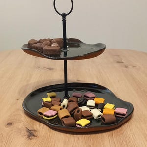 May include: A two-tiered serving tray with a black finish and a brown rim, filled with assorted candies and chocolates. The tray has a black metal handle and is set on a wooden surface.