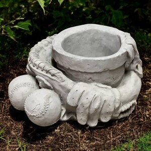 May include: A gray concrete baseball-themed planter. The planter is shaped like a baseball glove holding a pot, with two baseballs resting in front. The planter is designed for outdoor use and is suitable for planting flowers or small plants.