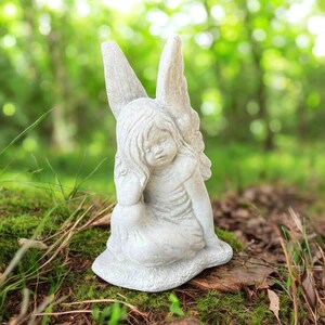 May include: A small, light gray angel statue with large, detailed wings. The angel is depicted in a seated position, with its head resting on its hand. The statue is made of a stone-like material and is suitable for garden or indoor decor.