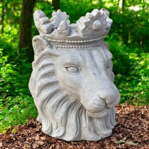 May include: A gray concrete lion head sculpture wearing a crown. The lion's face has detailed features, including a mane, eyes, and nose. The sculpture is outdoors, surrounded by greenery and mulch.
