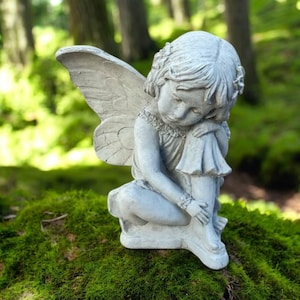 May include: A gray concrete fairy statue with detailed wings, a floral headpiece, and a seated pose. The fairy is resting her chin on her knee. The statue is set on a mossy surface, with a blurred forest background.