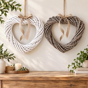 May include: Two heart-shaped wreaths made of woven branches, one painted white and one in a natural grey finish. Each wreath is suspended by a jute twine loop and features a decorative tan burlap ribbon bow tied at the top centre of the heart frame.