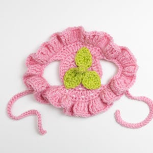 May include: A handmade pink crochet bonnet with a ruffled edge and green leaf accents. The bonnet has two pink ties and is designed for a baby or toddler. The bonnet is made of soft yarn and is likely comfortable to wear.
