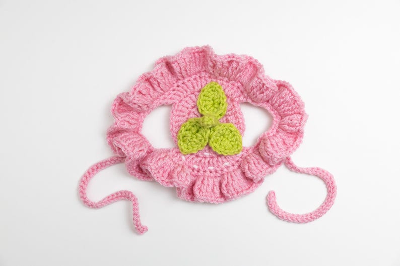 May include: A handmade pink crochet bonnet with a ruffled edge and green leaf accents. The bonnet has two pink ties and is designed for a baby or toddler. The bonnet is made of soft yarn and is likely comfortable to wear.