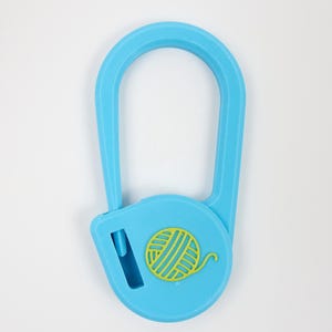 May include: A light blue yarn cutter pendant with a U-shaped loop and a circular base. The base features a yellow yarn ball icon and a vertical slit containing a blade for cutting yarn. The object has a textured finish and a simple design.