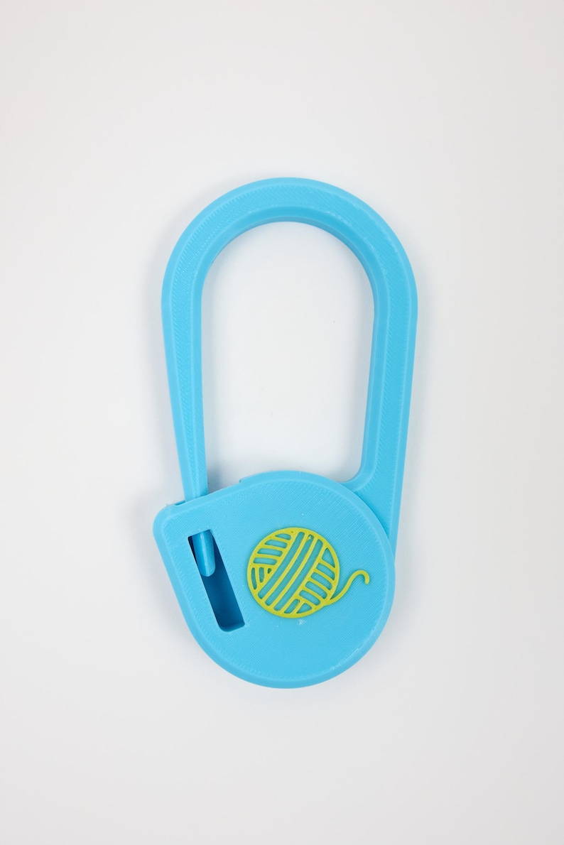 May include: A light blue yarn cutter pendant with a U-shaped loop and a circular base. The base features a yellow yarn ball icon and a vertical slit containing a blade for cutting yarn. The object has a textured finish and a simple design.