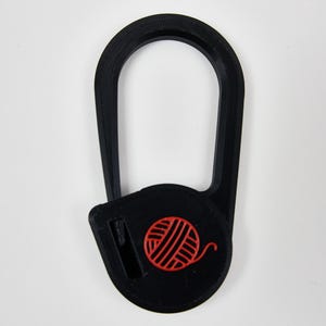 May include: A black plastic yarn cutter tool with an elongated oval shape and a circular base. The base features a bright red embossed icon of a ball of yarn and a small integrated blade slot for cutting yarn strands during knitting or crochet.