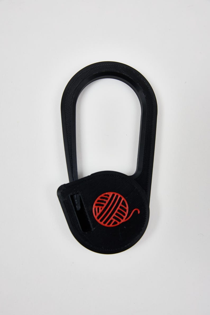 May include: A black plastic yarn cutter tool with an elongated oval shape and a circular base. The base features a bright red embossed icon of a ball of yarn and a small integrated blade slot for cutting yarn strands during knitting or crochet.