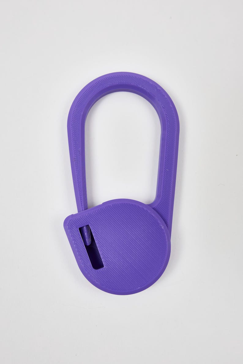 May include: A purple 3D printed plastic tool featuring a large oval handle attached to a circular base. The circular base contains a small rectangular cutout with a visible internal blade component, designed for cutting or slicing tasks.