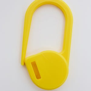 May include: A bright yellow plastic stitch marker shaped like a large safety pin. The object features a rounded base with a rectangular slot and a long, curved arm that extends upward and hooks into the base to create a secure, closed loop.