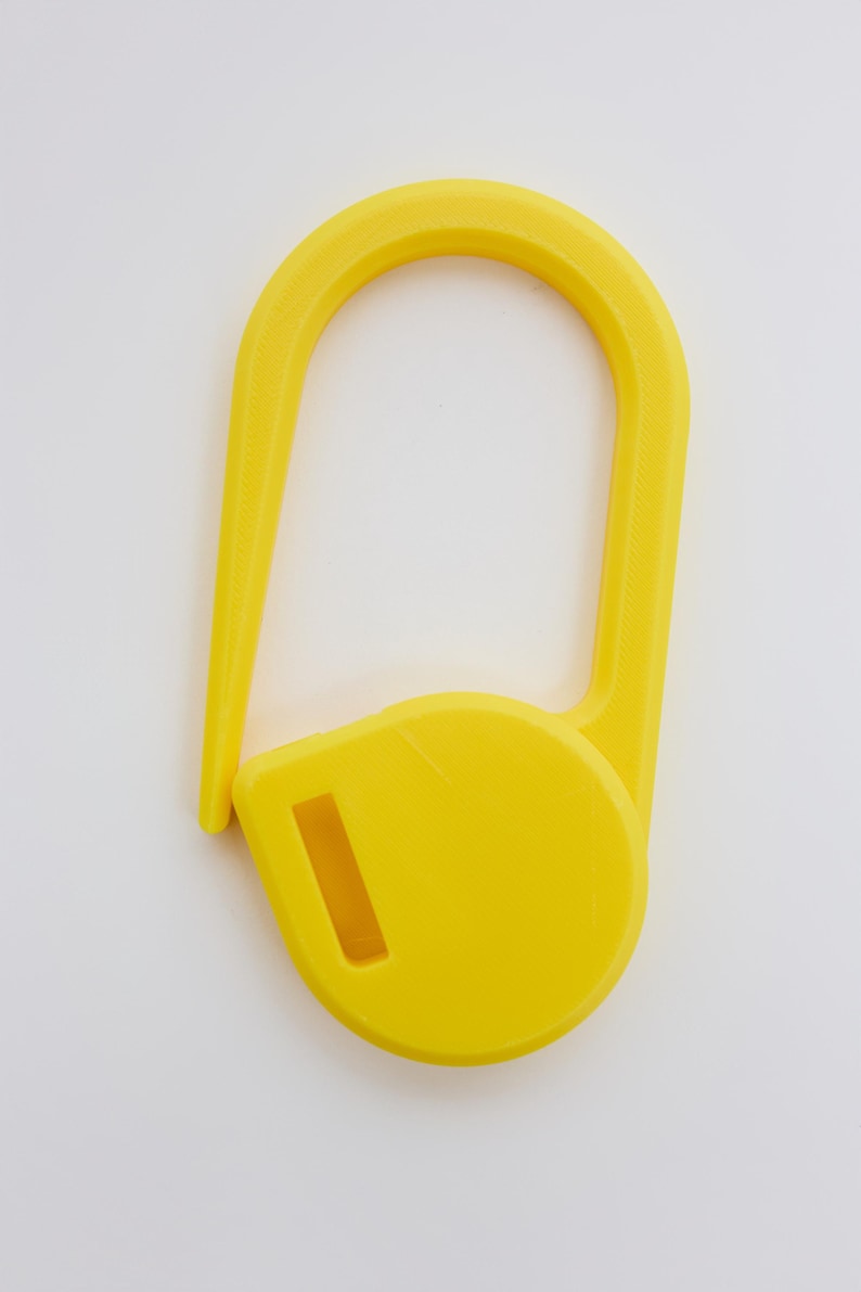 May include: A bright yellow plastic stitch marker shaped like a large safety pin. The object features a rounded base with a rectangular slot and a long, curved arm that extends upward and hooks into the base to create a secure, closed loop.