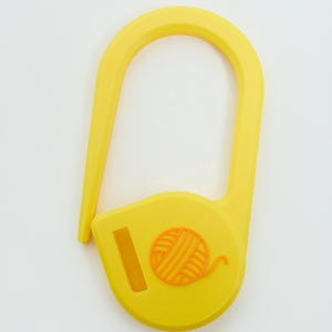 May include: A bright yellow plastic stitch marker shaped like a large safety pin. The base of the marker features a small, raised orange icon of a ball of yarn and a rectangular slot, providing a functional tool for knitting and crochet projects.