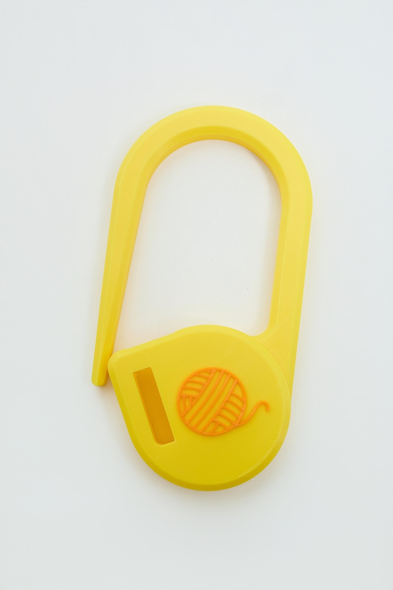 May include: A bright yellow plastic stitch marker shaped like a large safety pin. The base of the marker features a small, raised orange icon of a ball of yarn and a rectangular slot, providing a functional tool for knitting and crochet projects.