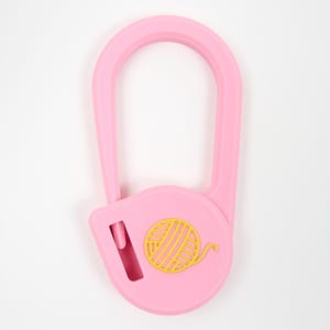 May include: A light pink plastic yarn cutter tool with a U-shaped handle and a circular base. The center of the circular base features a yellow embossed graphic of a ball of yarn. A small vertical blade slot is visible on the left side.