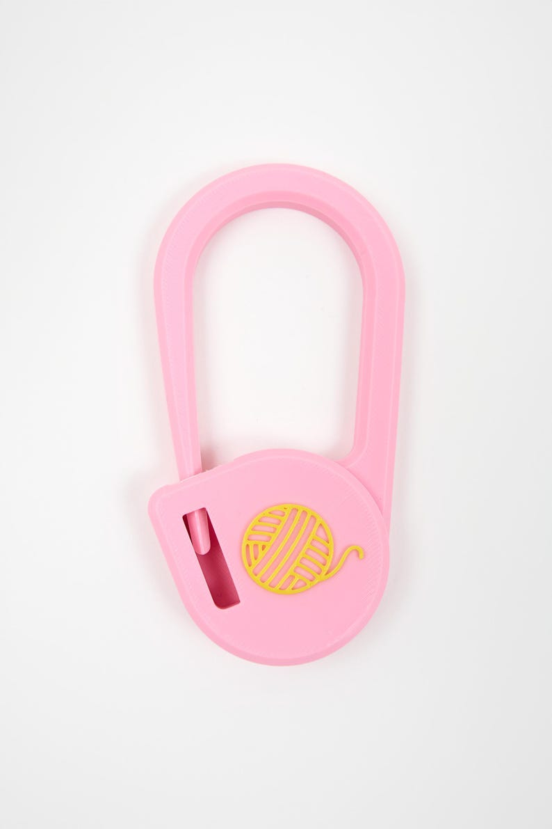 May include: A light pink plastic yarn cutter tool with a U-shaped handle and a circular base. The center of the circular base features a yellow embossed graphic of a ball of yarn. A small vertical blade slot is visible on the left side.