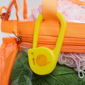 May include: A clear plastic storage bag with orange fabric trim and zippers, containing multiple skeins of yarn in pink, green, and red. A large, bright yellow plastic safety pin-shaped clip with a yarn ball icon hangs from the orange strap.