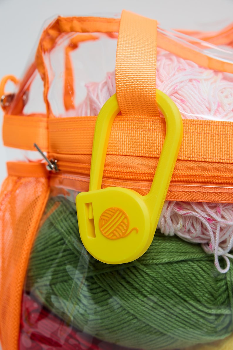 May include: A clear plastic storage bag with orange fabric trim and zippers, containing multiple skeins of yarn in pink, green, and red. A large, bright yellow plastic safety pin-shaped clip with a yarn ball icon hangs from the orange strap.