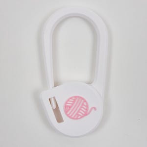 May include: A white plastic yarn cutter tool featuring a U-shaped frame and a circular base. The center of the circular base displays a raised, light pink graphic of a ball of yarn with a small blade slot positioned to the left of the design.