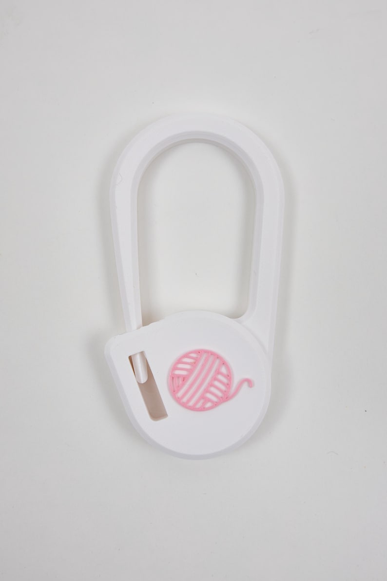 May include: A white plastic yarn cutter tool featuring a U-shaped frame and a circular base. The center of the circular base displays a raised, light pink graphic of a ball of yarn with a small blade slot positioned to the left of the design.