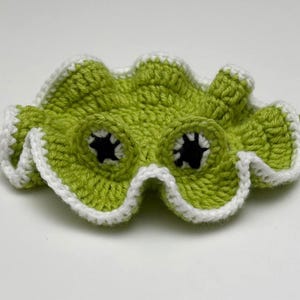 May include: A handmade, lime green crochet mask with white edging and black eye holes. The mask has a wavy, scalloped edge and two curved, green tendrils extending from the sides. The mask is designed to be worn.