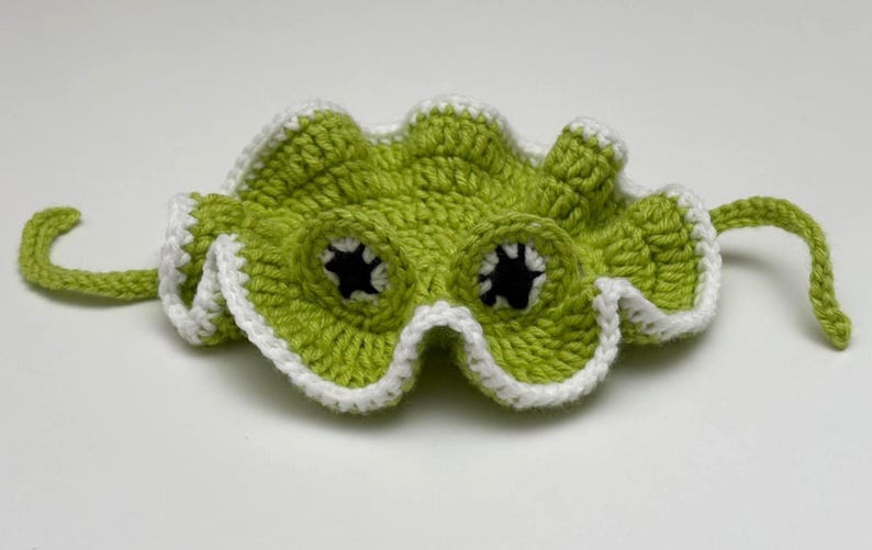 May include: A handmade, lime green crochet mask with white edging and black eye holes. The mask has a wavy, scalloped edge and two curved, green tendrils extending from the sides. The mask is designed to be worn.