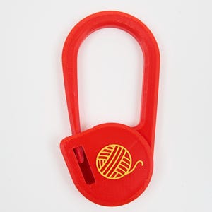 May include: A bright red plastic yarn cutter featuring a U-shaped handle and a circular base. The center of the circular base displays a yellow graphic icon of a ball of yarn. A small vertical blade slot is positioned on the left side of the base.