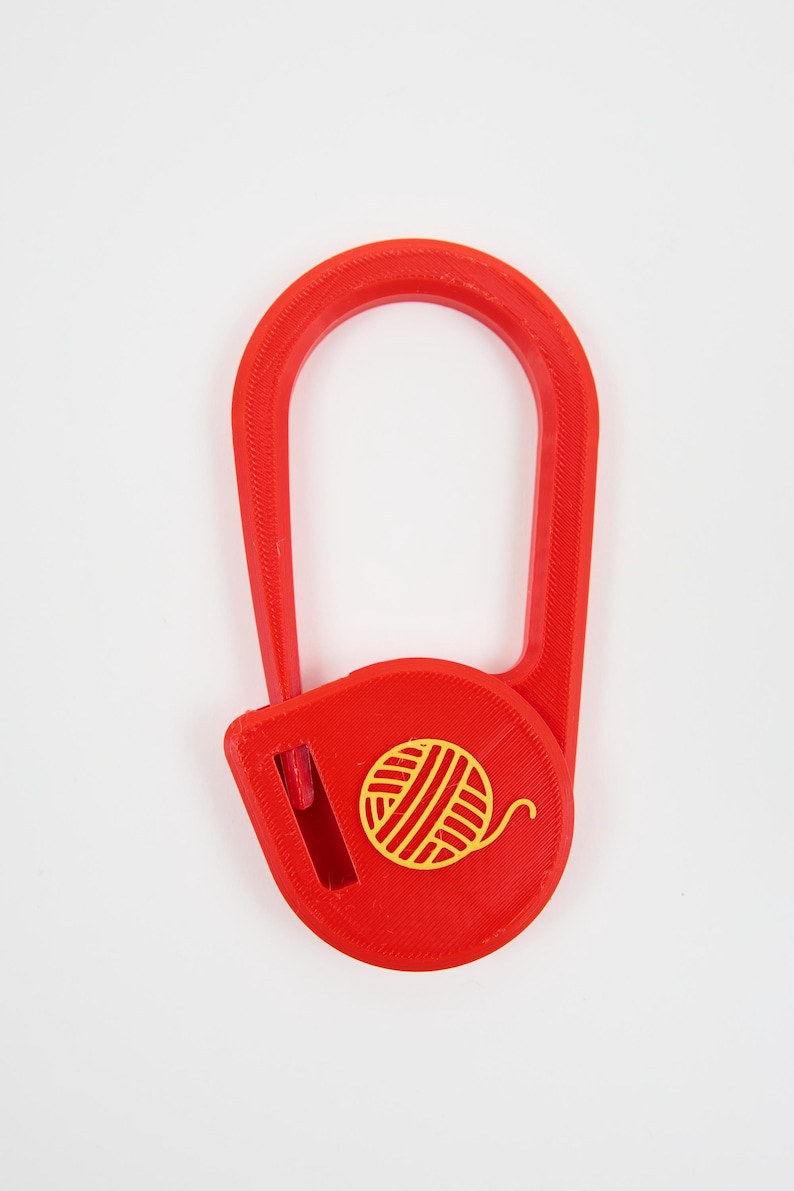 May include: A bright red plastic yarn cutter featuring a U-shaped handle and a circular base. The center of the circular base displays a yellow graphic icon of a ball of yarn. A small vertical blade slot is positioned on the left side of the base.