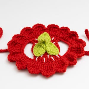 May include: A handmade, red crocheted flower with a scalloped edge and green leaves in the center. White thread accents the center. Two red crocheted ties extend from the sides. The craft item is set against a white background.
