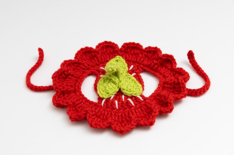 May include: A handmade, red crocheted flower with a scalloped edge and green leaves in the center. White thread accents the center. Two red crocheted ties extend from the sides. The craft item is set against a white background.