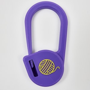 May include: A purple yarn cutter pendant with a teardrop-shaped frame and a circular base. The center of the circular base features a yellow yarn ball icon. A small rectangular slot on the left side of the base contains a metal blade for cutting.