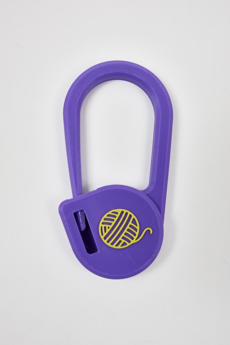 May include: A purple yarn cutter pendant with a teardrop-shaped frame and a circular base. The center of the circular base features a yellow yarn ball icon. A small rectangular slot on the left side of the base contains a metal blade for cutting.