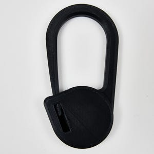 May include: A black 3D printed object featuring a large, elongated oval loop connected to a circular base. The circular base contains a small, rectangular slot positioned off-center. The entire piece has a visible layered texture.