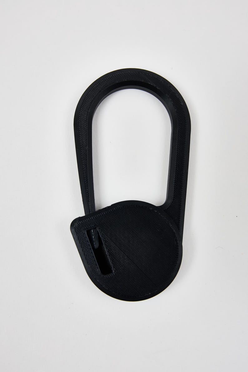 May include: A black 3D printed object featuring a large, elongated oval loop connected to a circular base. The circular base contains a small, rectangular slot positioned off-center. The entire piece has a visible layered texture.