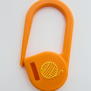 May include: A bright orange yarn guide tool featuring a U-shaped hook and a circular base. The base displays a raised yellow icon of a ball of yarn and includes a rectangular slot for threading yarn through during knitting or crochet projects.
