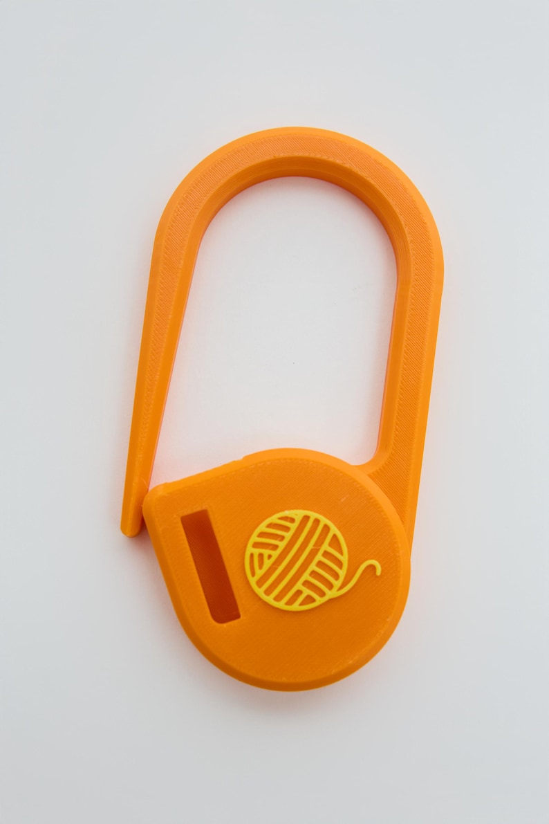 May include: A bright orange yarn guide tool featuring a U-shaped hook and a circular base. The base displays a raised yellow icon of a ball of yarn and includes a rectangular slot for threading yarn through during knitting or crochet projects.