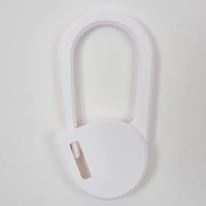 May include: A white plastic carabiner-style clip featuring a smooth, rounded oval frame and a circular locking mechanism at the base. The design includes a small, integrated rectangular slot on the lower circular section, providing a clean, solid white finish.