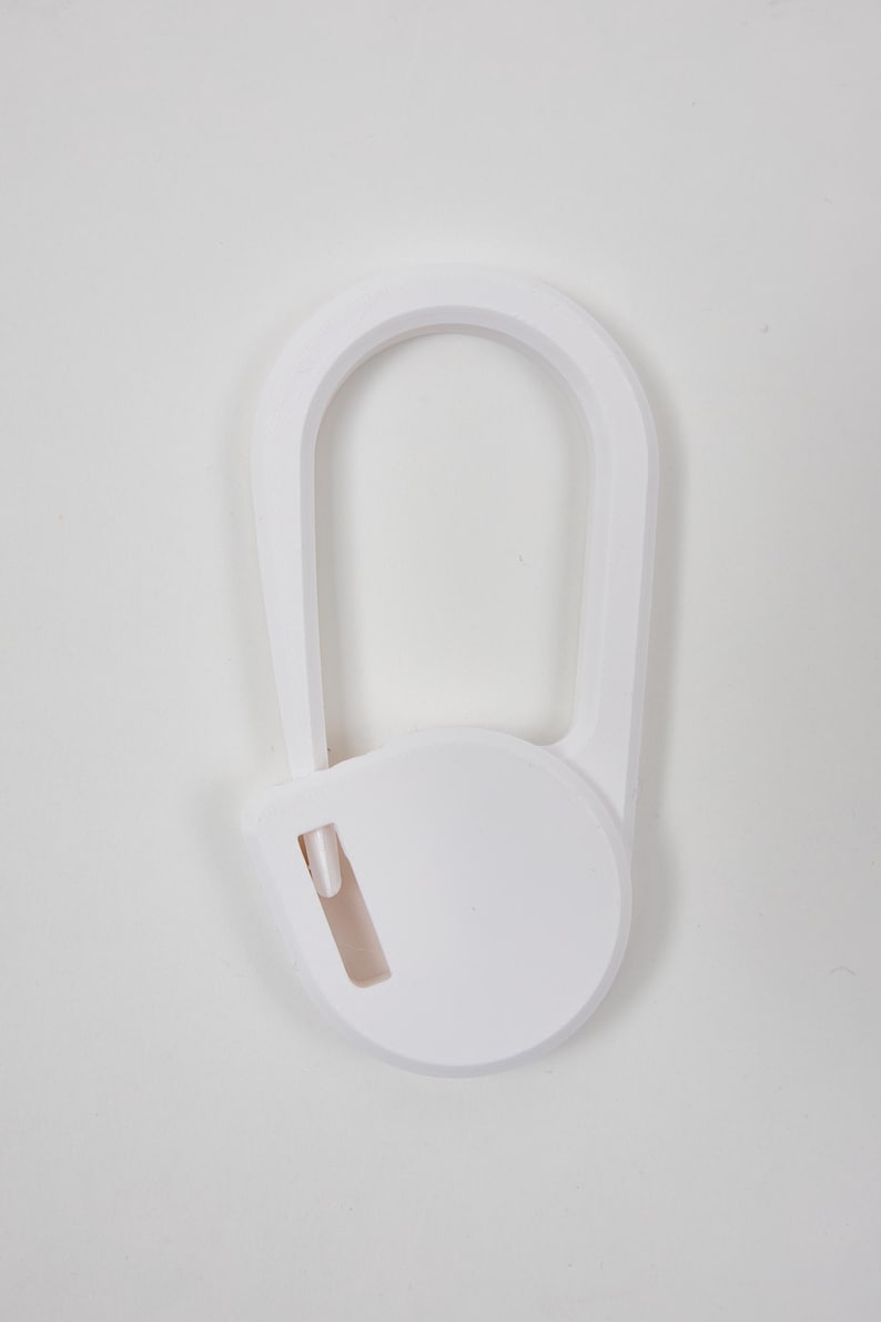 May include: A white plastic carabiner-style clip featuring a smooth, rounded oval frame and a circular locking mechanism at the base. The design includes a small, integrated rectangular slot on the lower circular section, providing a clean, solid white finish.