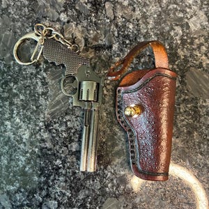 Puede incluir: Un llavero de revólver plateado con una funda de cuero en miniatura a juego. La funda es de color marrón oscuro con un botón dorado. El llavero tiene un anillo y una cadena para sujetarlo a llaves o a un bolso.